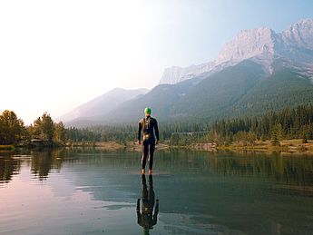 Leistungsdiagnostik, Laktatstufentest, Laktattest, Belastungstest, Kapillaren, Kapillarblut Entnahme. Ein Triathlon Sportler im Wasser vor einem Berg in den Alpen mit Neopren Anzug vor dem Schwimmen unter Beobachtung vom Trainer und Coach zur Verbesserung der Schwimmleistung und Vorbereitung zum Outdoor Abenteuer Wettkampf im Allgäu